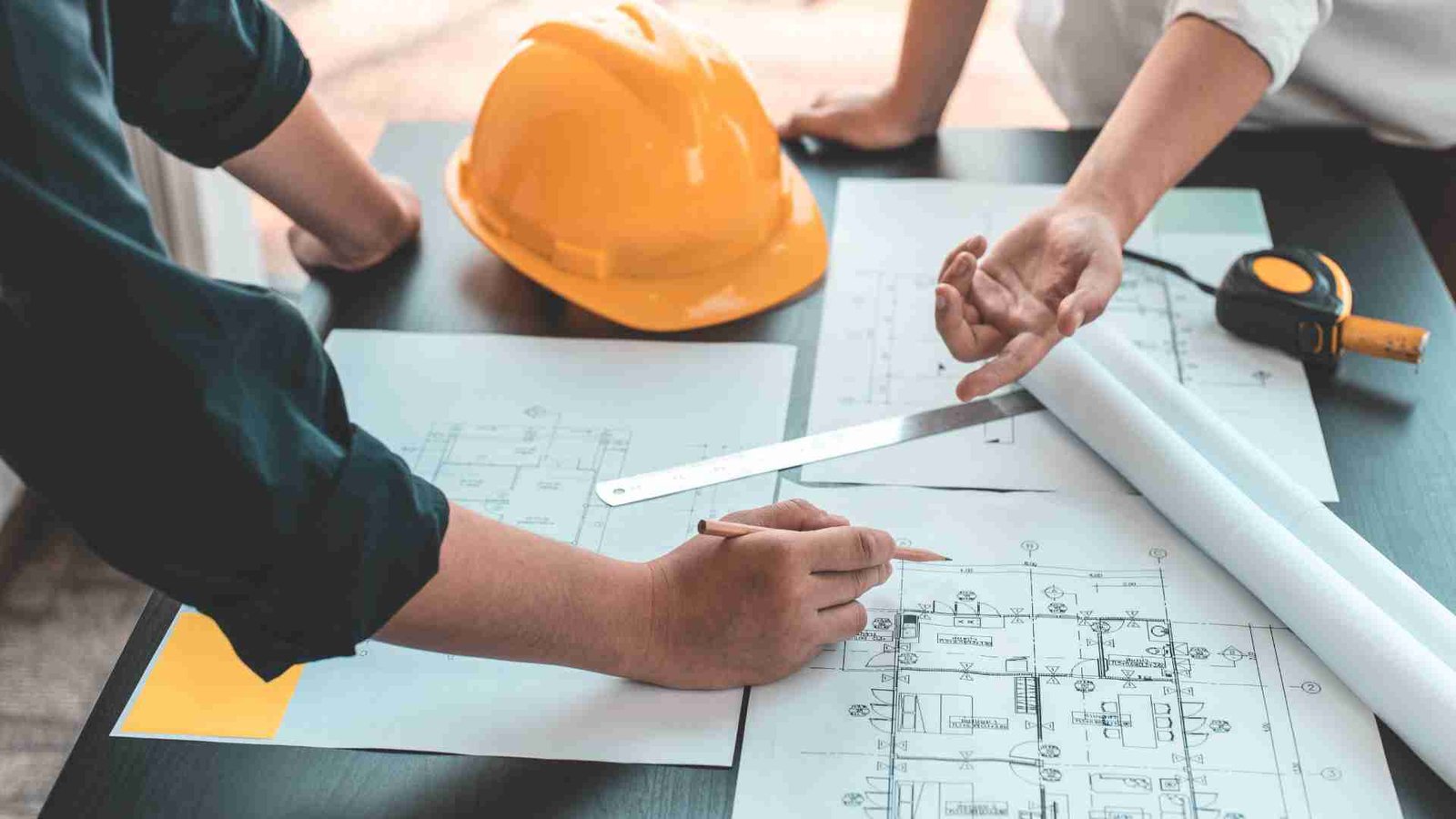 Illustrative building plans being reviewed beside a hard hat and measuring tools